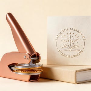 May include: A rose gold library seal next to a cream-colored card and a book. The card is embossed with the text "FROM THE LIBRARY OF HANNAH ROBERTS" around a design of an open book, a cross, and stars. The seal is open, ready to be used.