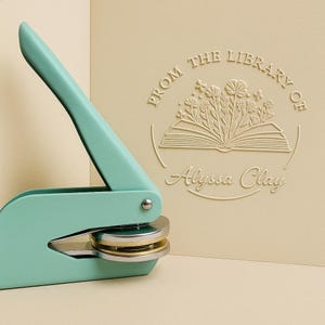 May include: A teal library seal with a silver and gold die is positioned next to a cream-colored card. The card features embossed text that reads "From the Library of Alyssa Clay" with a floral book design.
