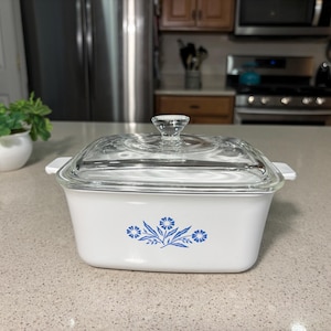 May include: A white rectangular casserole dish with a clear glass lid. The dish features a blue floral design on the front. The lid has a clear glass knob. The dish is on a light-colored countertop.