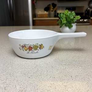 Könnte beinhalten: Ein weißer Keramik-Kochtopf mit Griff. Der Kochtopf hat ein Blumenmuster mit einer roten Paprika, einer grünen Artischocke und einer gelben Zitrone. Der Text "Le Persil" ist auf dem Boden des Topfes aufgedruckt.