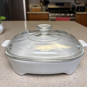 May include: A white, square casserole dish with a clear glass lid. The dish has two white handles and is on a speckled countertop. The lid has a clear glass knob. The dish is likely used for baking or serving food.