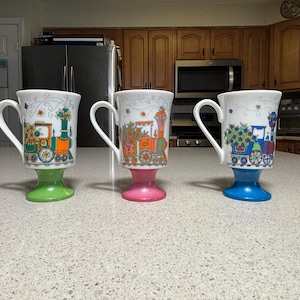 May include: Three white footed mugs with a train design. Each mug has a different colored base: green, pink, and blue. The train design features orange, green, and blue colors with floral accents. The mugs have a curved handle.