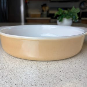 May include: A light beige round baking dish with two handles. The dish is made of glass and has a white interior.