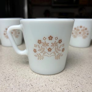 May include: Three white ceramic mugs with brown floral designs. The mugs have a simple, rounded shape and a small handle.