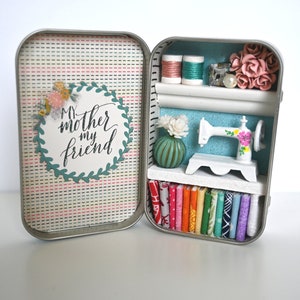 May include: Open tin box diorama with a sewing theme. The left side has a patterned background with the text "My mother my friend". The right side has a shelf with spools of thread, a miniature sewing machine, and fabric samples. Colors include pink, green, and white.