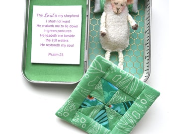 Könnte beinhalten: Eine handgefertigte Miniatur-Szene mit einem schlafenden Schaf in einer grünen Dose. Die Dose enthält ein kleines Kissen, ein Bett und eine Karte mit dem Text "The Lord is my shepherd". Ein grün-blaues, gestepptes Quadrat liegt neben der Dose.
