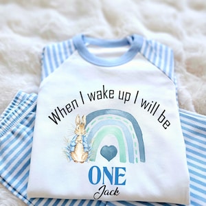 May include: White onesie with blue stripes on the sleeves and legs. The onesie has a Peter Rabbit illustration, a rainbow, and the text "When I wake up I will be one Jack".
