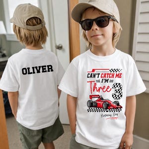 May include: Two white t-shirts with black text and graphics. One shirt has the name "OLIVER" on the back. The other shirt has a race car graphic and the text "CAN'T CATCH ME I'M Three" with a number 3 and "Birthday Boy".