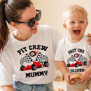 Op de afbeelding: Witte T-shirts met bijpassende raceauto-ontwerpen. Eén shirt leest "PIT CREW MUMMY" met een rode raceauto en geblokte vlaggen. Het andere shirt zegt "FAST ONE OLIVER" met een vergelijkbaar ontwerp. Moeder en kind dragen de shirts.