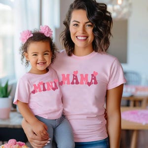 May include: A pink t-shirt with the text "Mini Mama" in pink and red letters with pink hearts. A child is wearing a matching pink t-shirt with the text "Mini" in pink and red letters with pink hearts.