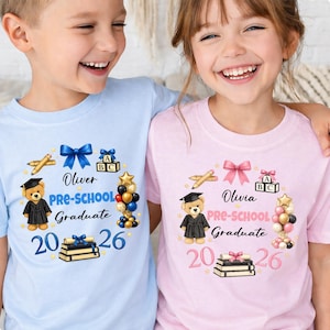 May include: Two children wearing light blue and pink preschool graduation t-shirts. The shirts feature a teddy bear in a graduation gown, pencils, balloons, and the text "Oliver PRE-SCHOOL Graduate 2026" and "Olivia PRE-SCHOOL Graduate 2026".