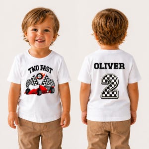 May include: White t-shirt featuring a red race car graphic with black and white checkered flags and the text "TWO FAST". The back of the shirt displays the name "OLIVER" and a checkered number "2".