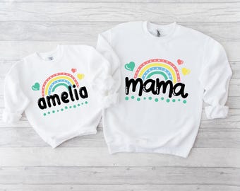 May include: Two white sweatshirts with rainbow and heart designs. The larger sweatshirt says "mama" and the smaller sweatshirt says "amelia".