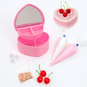 Può includere: Un portagioie rosa a forma di cuore con uno specchio, aperto per rivelare scomparti. Una piccola torta rosa a forma di cuore con ciliegie, sac à poche, zuccherini e ciliegie sono anche presenti.