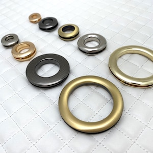 May include: Assortment of metal grommets in various sizes and finishes, including gold, silver, and black. The grommets are arranged in a row on a white quilted surface.