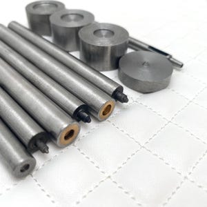 May include: A set of metal tools for leather crafting, including a punch, a hammer, and several different sized dies. The tools are arranged on a white surface.