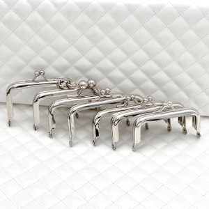 Puede incluir: Un conjunto de ocho marcos de bolso plateados con bordes redondeados y cierre en forma de bola. Los marcos están dispuestos en fila, con el marco más grande en la parte superior y el marco más pequeño en la parte inferior.