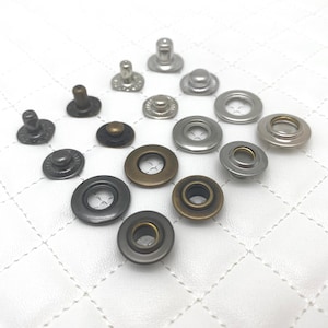 Puede incluir: Surtido de botones a presión metálicos de varios tamaños y acabados, incluyendo plata, latón y negro. Los botones a presión están dispuestos sobre una superficie blanca.