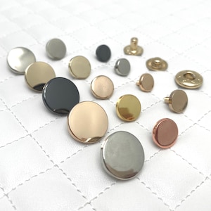 Puede incluir: Surtido de botones redondos de metal en varios acabados, incluyendo plata, oro, oro rosa, cobre y negro. Los botones se muestran sobre una superficie acolchada blanca.