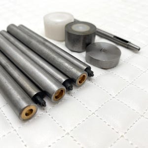 Könnte beinhalten: Ein Satz Metallwerkzeuge zum Setzen von Ledernieten. Das Set enthält einen Stempel, einen Hammer und mehrere Nietensetzer in verschiedenen Größen. Die Werkzeuge liegen auf einer weißen Oberfläche.