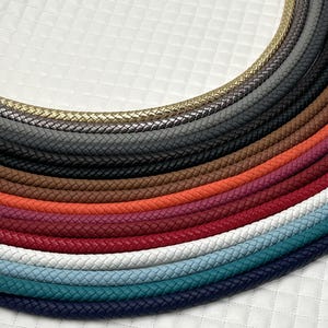 May include: A selection of braided leather cords in a range of colours, such as gold, grey, black, brown, red, white, teal, and navy. The cords are arranged in a semi-circular pattern against a white, quilted background.