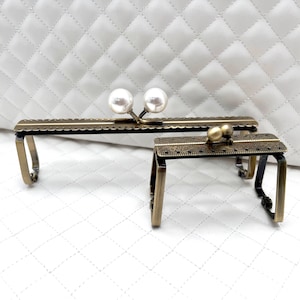 Puede incluir: Dos marcos de bolso de metal dorado antiguo con detalles decorativos. El marco superior tiene dos cuentas blancas perladas. El marco inferior tiene una sola cuenta redonda.