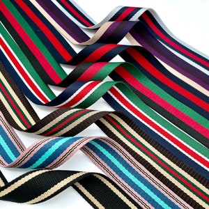 Könnte beinhalten: Sortiment an gestreiften Bandbesätzen in verschiedenen Farben, darunter Schwarz, Weiß, Rot, Grün, Blau und Beige. Die Bänder sind mit unterschiedlich farbigen Streifen gewebt und in einem mehrschichtigen, überlappenden Muster angeordnet.