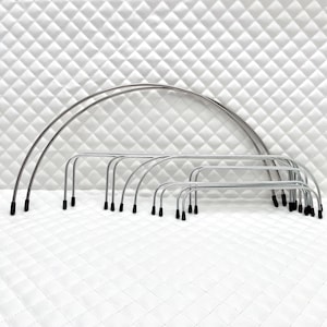 May include: A collection of silver-colored metal bra underwires in various sizes, each with black tips. The underwires are arranged in a row, showcasing their different widths and curved shapes. The background is a white, quilted fabric.