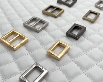 May include: Assortment of metal slider buckles in various finishes, including gold, silver, and black. The buckles are rectangular with a center bar and are arranged on a white quilted surface.