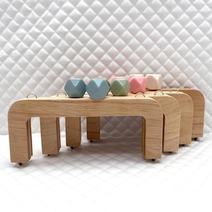 Può includere: Quattro manici in legno con anelli metallici, impilati uno sopra l'altro. I manici sono progettati per essere utilizzati per trasportare borse o sacche. Cinque forme geometriche colorate poggiano sui manici.