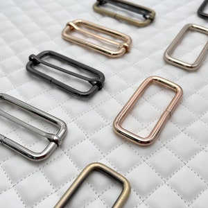 May include: Assortment of metal slider buckles in various finishes, including silver, gold, and black. The buckles are rectangular in shape and have a smooth, polished finish.