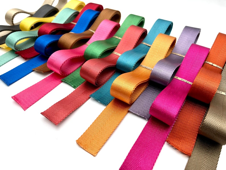 May include: Assortment of colourful woven ribbon in various shades of green, blue, red, orange, yellow, and brown. The ribbon is arranged in a fan shape on a white background.