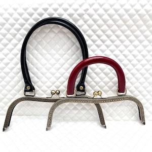Op de afbeelding: Twee metalen tasframes met decoratieve details. Een frame is bevestigd aan een zwart leren handvat en de andere aan een rood leren handvat.