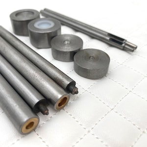 May include: A collection of metal tools, including cylindrical punches and rods, arranged on a white, quilted surface. The tools vary in size and shape, with some having rounded ends and others featuring hollow interiors.