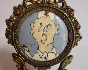 May include: A brass picture frame with a small round portrait of a person wearing a white hat with a red cross. The frame has a decorative design and stands on a small easel.