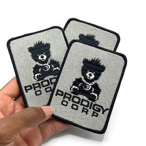 May include: Three gray embroidered patches with a black border. Each patch features a black teddy bear wearing a chain and the text "PRODIGY CORP" in black. The patches are rectangular with rounded corners.