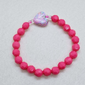 Puede incluir: Un brazalete de cuentas rosadas con un dije en forma de corazón. El dije es rosa, morado y azul con un patrón floral.