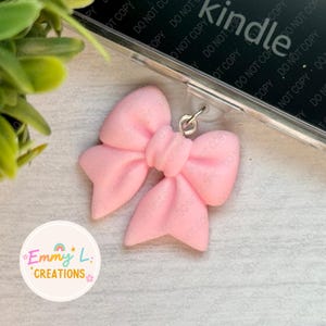 May include: A light pink bow charm with a silver-colored clasp. The bow is made of a soft material and is attached to a clear phone case. The charm is a decorative accessory. The image also includes the text "Emmy L. Creations".