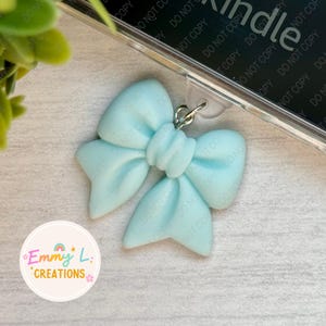 May include: A light blue bow charm with a silver ring and a clear plastic phone case with the word "Kindle" printed on it. The charm is attached to the phone case with a clear plastic ring.