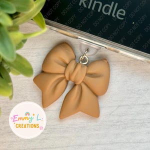 May include: A tan-colored bow-shaped badge reel charm. The charm is made of a smooth material and has a silver-colored metal clasp. The text "Emmy L. Creations" is printed on a white circular sticker.