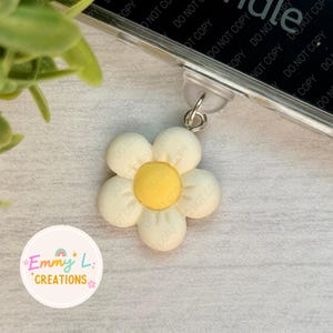 May include: A white and yellow daisy charm with a silver ring attached. The charm is hanging from a clear phone case.