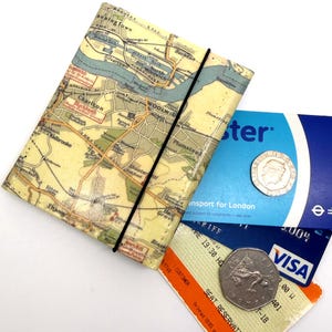 May include: A rectangular wallet featuring a vintage map print of London, secured with a thin black elastic band. Inside are a blue Transport for London card, a blue Visa debit card, an orange seat reservation ticket, and two silver coins.