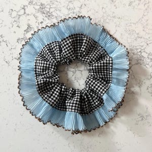 May include: A scrunchie with a black and white gingham pattern center, surrounded by a light blue pleated fabric and a brown trim. This hair accessory is a stylish addition to any hairstyle.