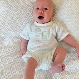 May include: A baby wearing a white romper with the name "Cal" embroidered in light green. The romper has short sleeves and a textured pattern. The baby is lying on a white, textured blanket.