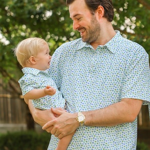 May include: A father and child wearing matching short-sleeved shirts with a light blue and green floral pattern. The father is holding the child, both smiling. The father is wearing a gold watch.