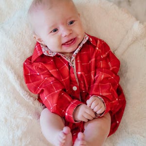 May include: A smiling baby wearing a red plaid shirt with a white and patterned collar. The baby is lying on a soft, white, fluffy blanket. The shirt has a single white button.