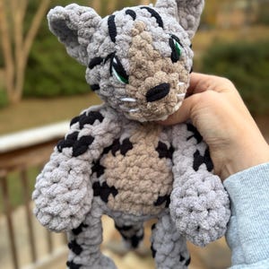 May include: A handmade crochet cat plushie in gray with black spots and stripes. The cat has a tan belly, green eyes, and a black nose. The plushie is held in a person's hand.