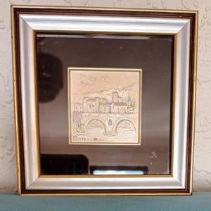Puede incluir: Una obra de arte en relieve de plata enmarcada que representa un puente y edificios en un paisaje. La obra de arte est&aacute; en un marco marr&oacute;n y plateado con un passe-partout negro. El texto "R 1+20" es visible en la esquina inferior izquierda de la obra de arte.