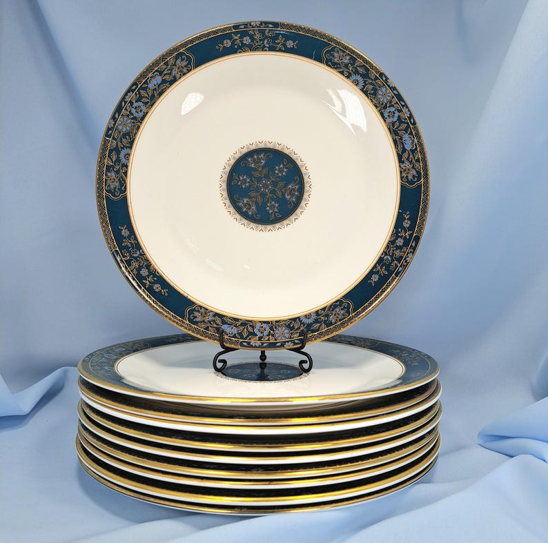 Op de afbeelding: Een stapel van acht ronde keramische borden met een cr&egrave;mekleurig midden, een brede donkerblauwe rand met gedetailleerd goud filigraan en een centraal rond medaillon met blauwe en gouden bloemmotieven en goudkleurige randen.