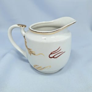 May include: White ceramic creamer with a curved handle and spout. The creamer has gold trim and decorative gold and brown leaf-like designs. The handle has a gold accent. The creamer is on a light blue surface.